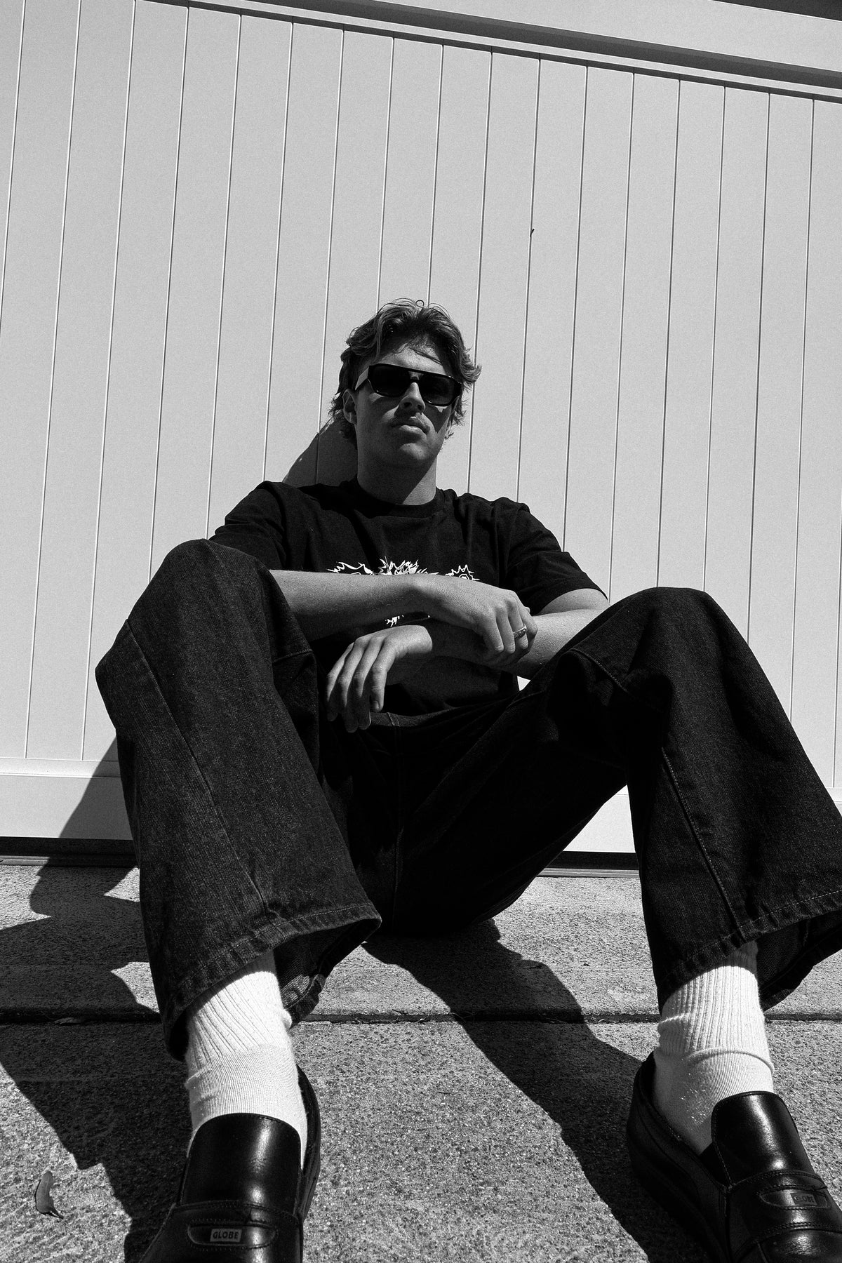 A black and white lifestyle photograph of a man sitting on concrete in front of a white paneled wall. He is wearing dark sunglasses, a black graphic t-shirt, dark wide-leg jeans, white crew socks, and black leather loafers.
