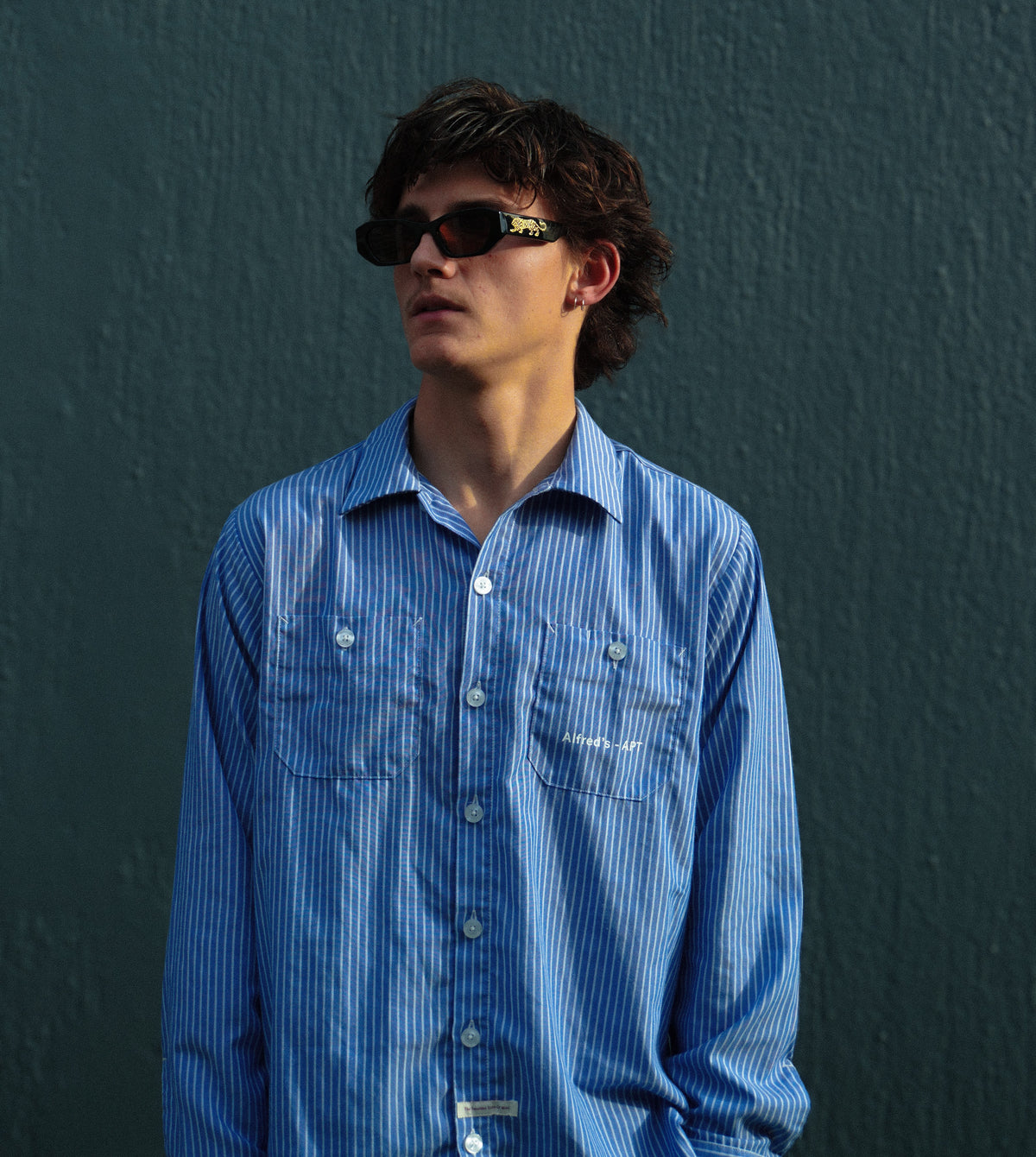 A man wearing a blue and white vertically striped button-down shirt with chest pockets, standing against a dark textured wall.